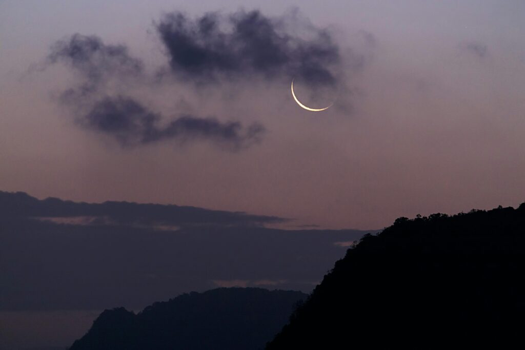 The new moon, which signals the beginning of a new Islamic month, including Ramadan and Shawwal, on a dusty purple night sky above forest mountains